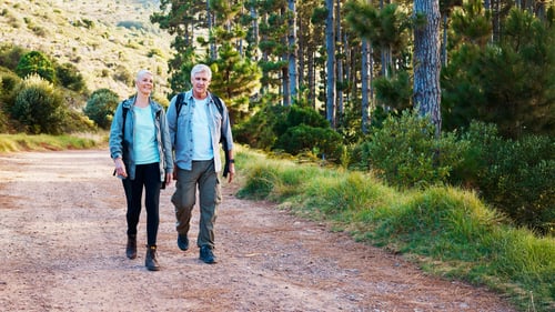 Two senior hikers walking together on a dirt trail through a scenic landscape with pine trees on the right and a hillside on the left. The setting appears to be a beautiful nature trail in late afternoon light.