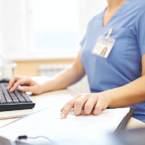 Healthcare worker in blue scrubs with ID badge typing on computer keyboard while reviewing documents at desk in medical office setting.