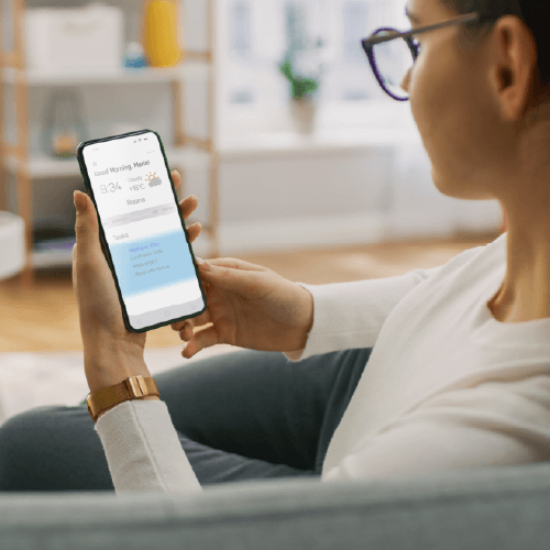 Woman wearing glasses and a white sweater sits on a couch using a smartphone, viewing what appears to be a healthcare or medical app interface. She's wearing a gold watch and the setting is a bright, modern living room with shelving visible in the background.