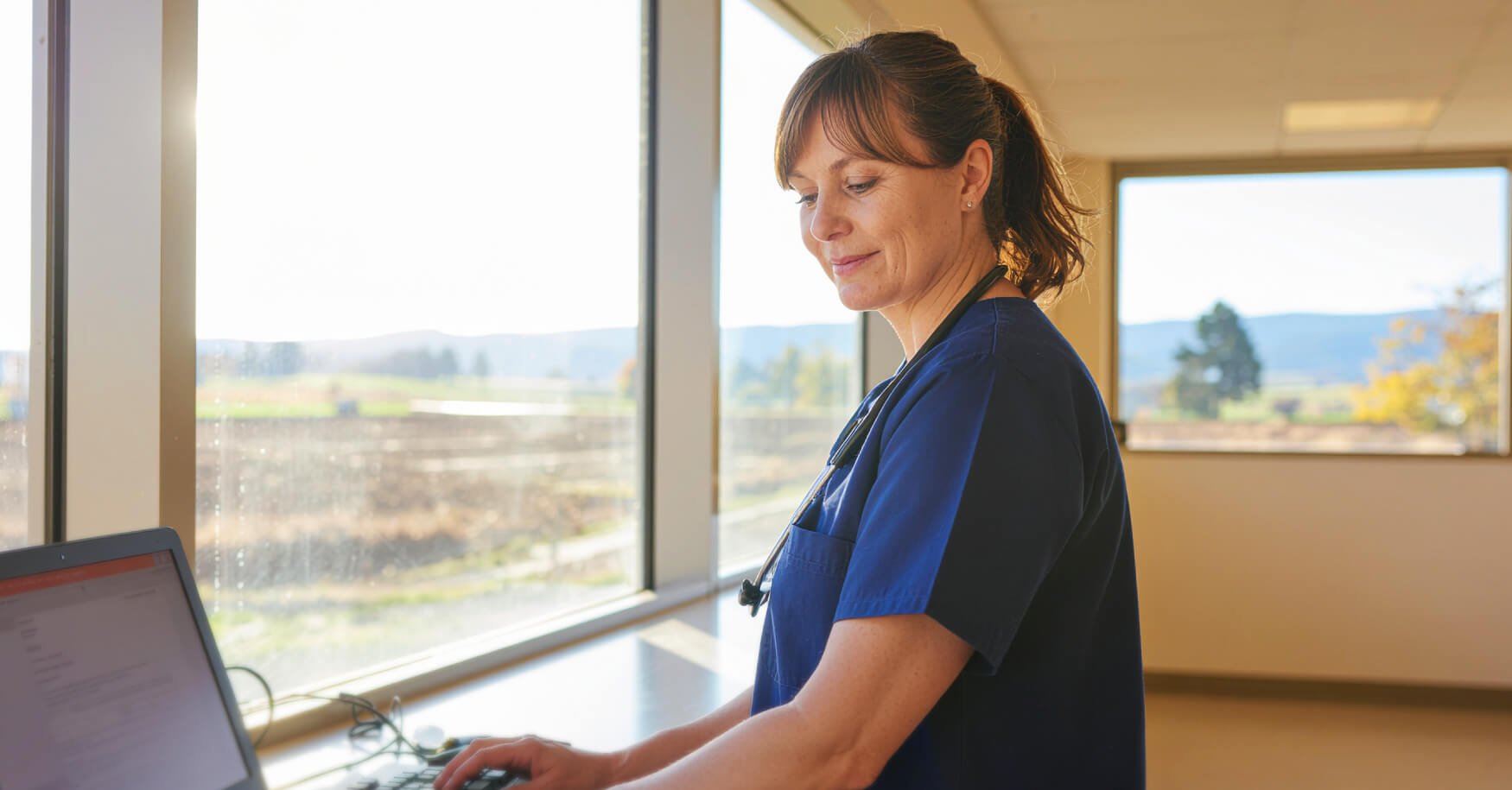 Female nurse in blue scrubs with stethoscope working at a computer workstation in a rural hospital facility