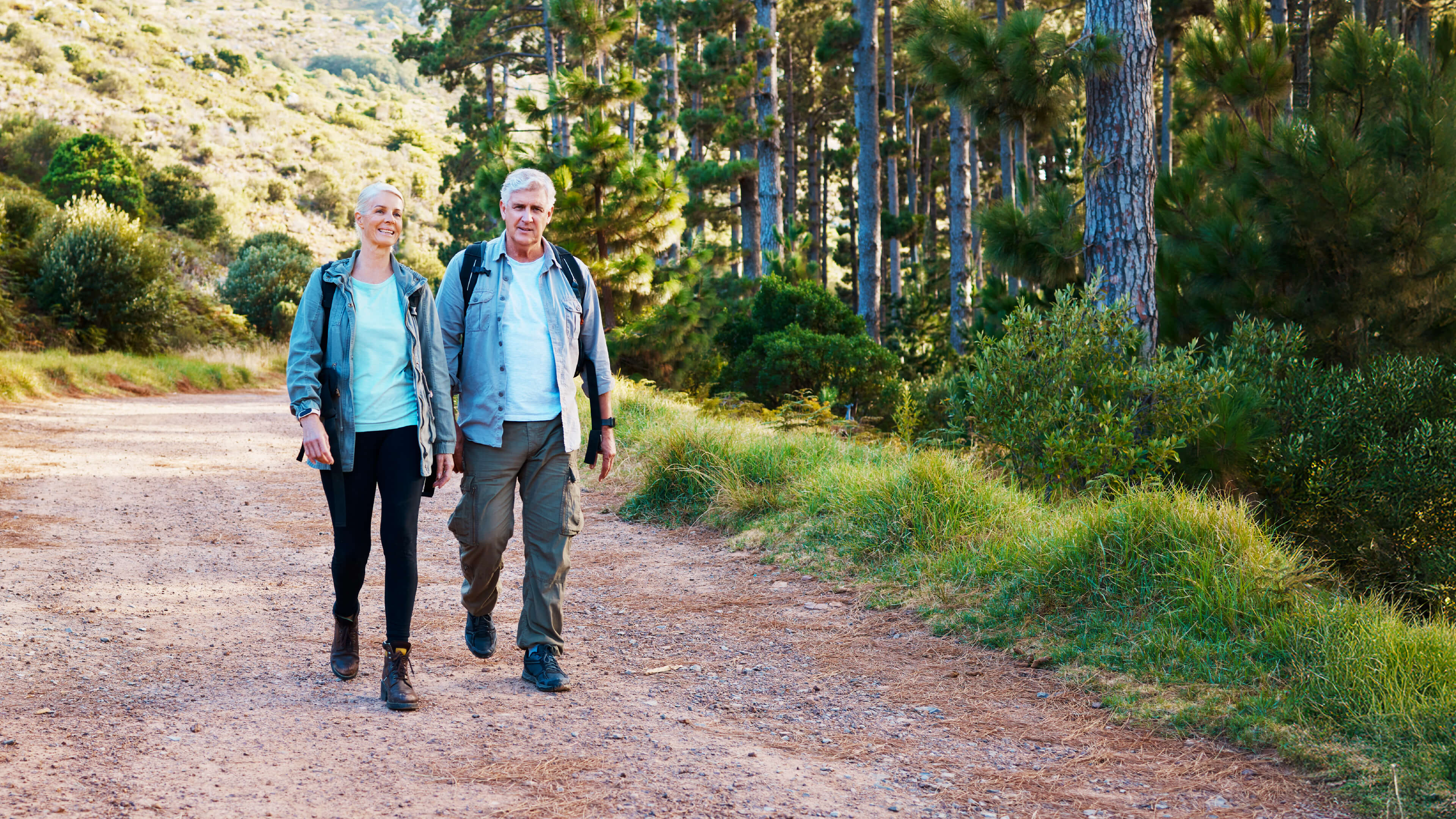 Two senior hikers walking together on a dirt trail through a scenic landscape with pine trees on the right and a hillside on the left. The setting appears to be a beautiful nature trail in late afternoon light.