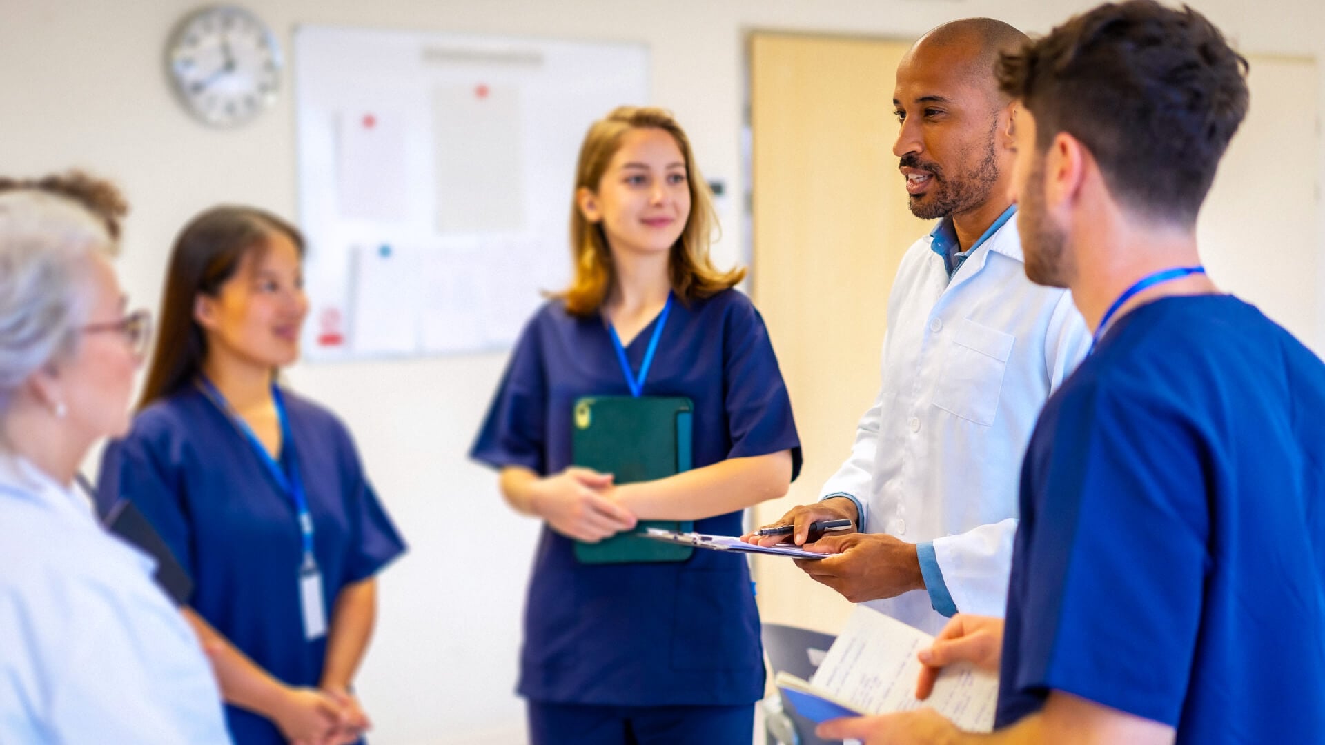 A diverse team of behavioral health clinicians collaborating during a care team huddle in a clinical setting, with staff in scrubs and a white coat reviewing patient information.