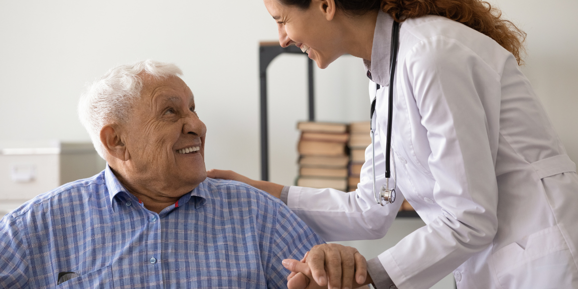 A smiling female doctor in a white coat and stethoscope holds the hand of an elderly male patient during a checkup, reflecting the personalized care found in rural healthcare settings.